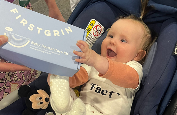 FirstGrinBaby – CareQuest Innovation Partners Baby in a stroller reaching for a light blue box labeled 'Baby Dental Care Kit' held by a caregiver side.