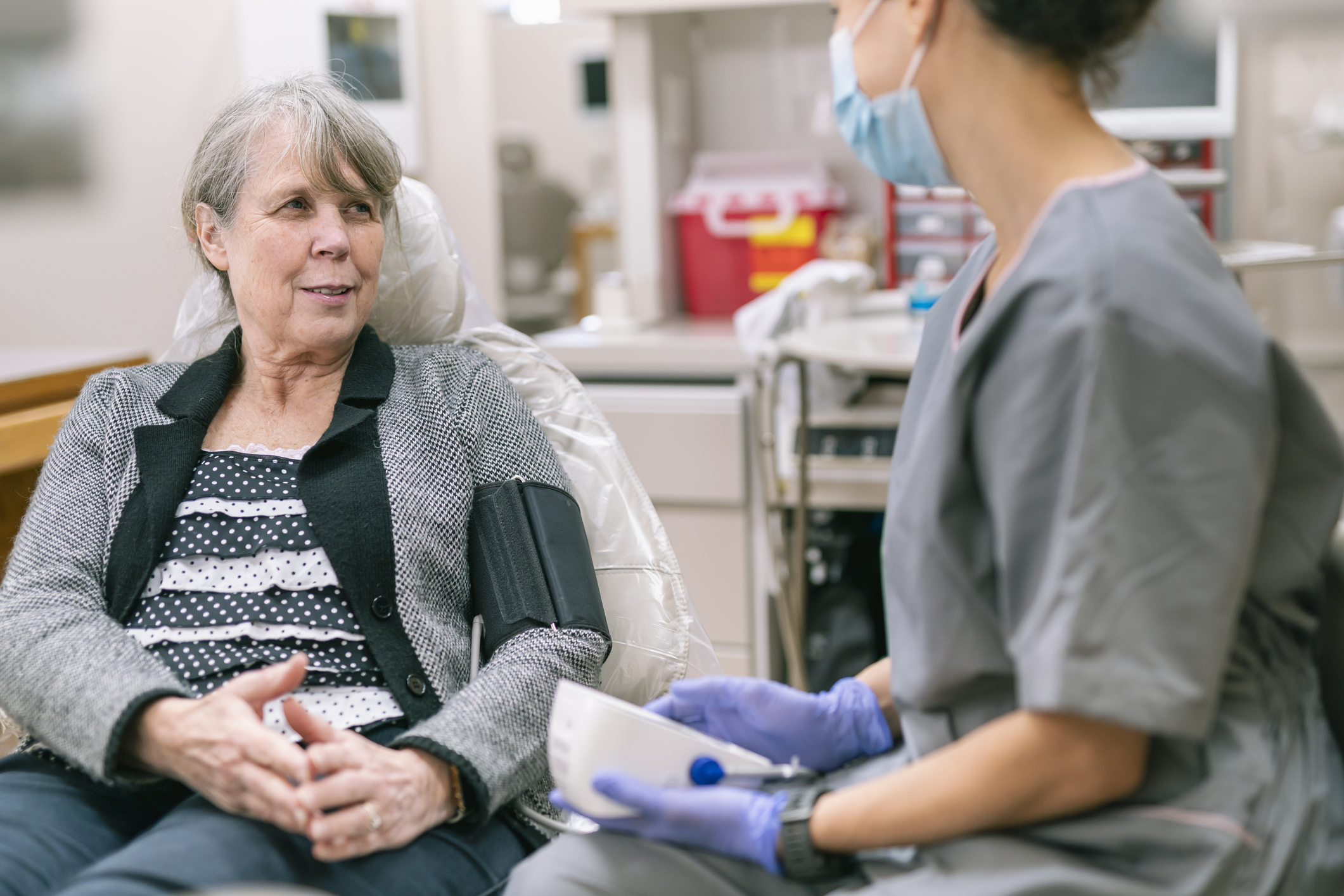 Elderly woman sits in a chair as a nurse in scrubs and a mask prepares a medical procedure nearby in a clinic room (gloved hands visible).