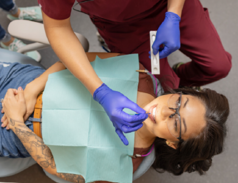 Dentist in purple scrubs with blue gloves treats a patient reclining in a dental chair, using a dental instrument near the mouth.