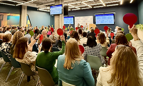 Demo Day 1 – CareQuest Innovation Partners Audience in a conference room raising red circular signs while a panel speaks at the front with multiple screens displaying slides.