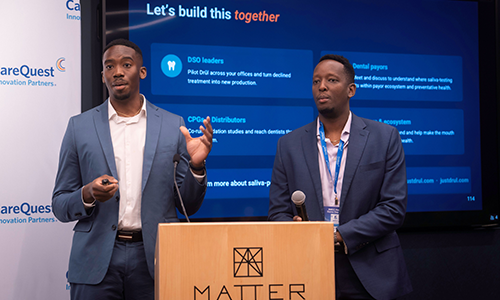 Drul Pitch – CareQuest Innovation Partners Two men in blue suits present on a stage, standing behind a wooden MATTER podium with a large screen behind them.