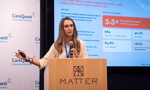 Firstgrin Pitch – CareQuest Innovation Partners Woman presenting at a MATTER podium with microphone, gesturing with her left hand as a slide with data appears to the right; CareQuest branding in the background.