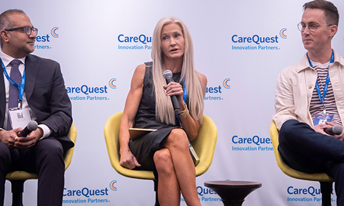 Interop Panel – CareQuest Innovation Partners Three panelists seated in yellow chairs on a stage with CareQuest logos behind them; the center woman speaks into a handheld microphone while a man to her left holds another mic and a man to her right listens.