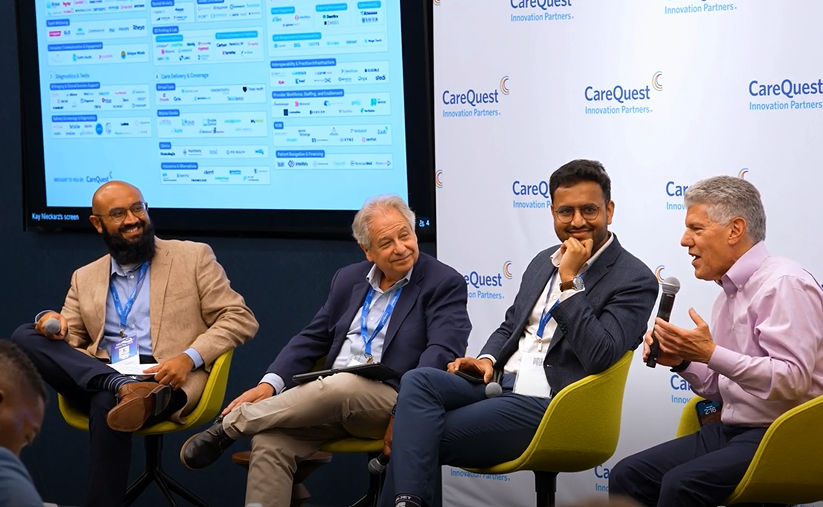 Four men in a panel discussion seated on yellow chairs, one speaking into a handheld microphone against a CareQuest backdrop with blue lanyards and name badges.
