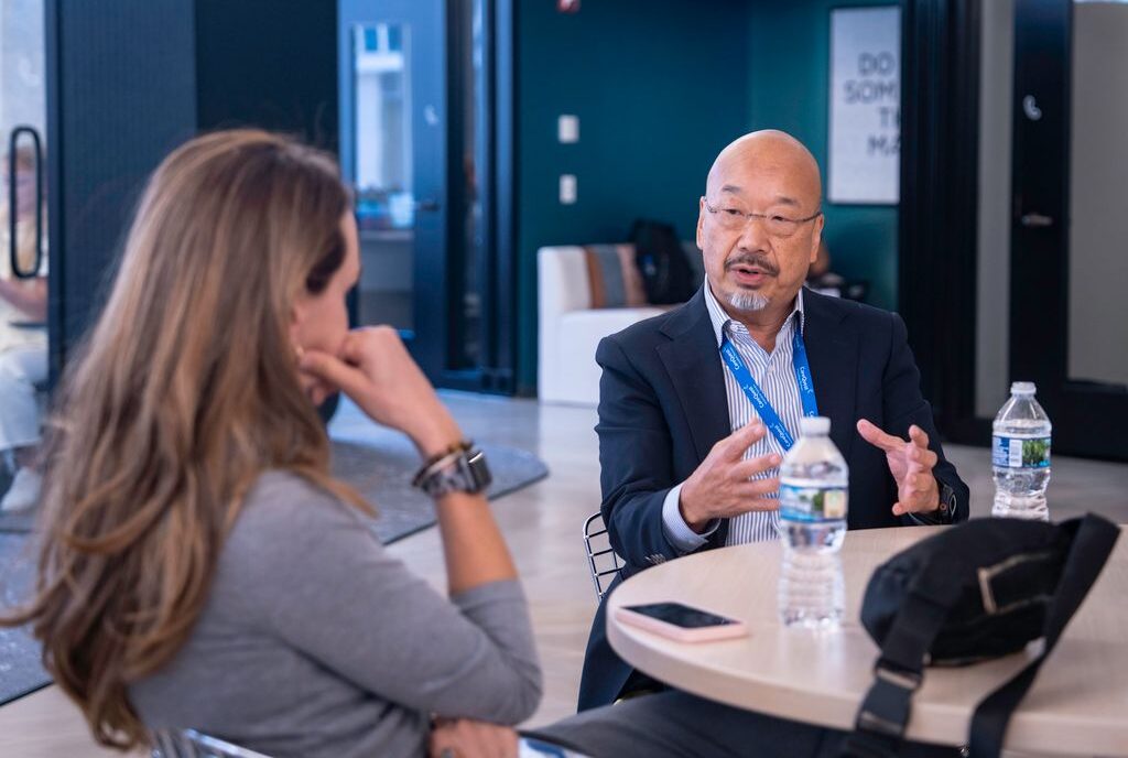 Two professionals sit at a round table in a modern office; a bald Asian man in a suit explains something with his hands while a woman with long hair listens across from him.