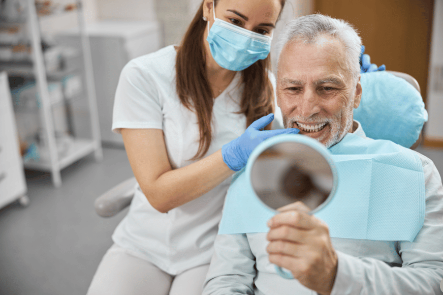 Dental assistant in a mask and gloves helps an elderly man in a dental chair, as he looks at his reflection in a handheld mirror.