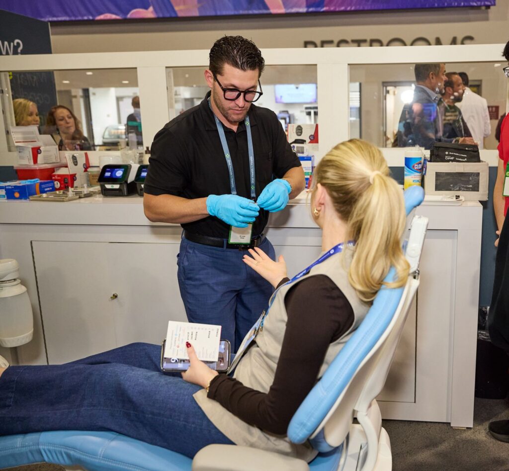 Dentist in a black shirt and blue gloves works on a patient seated in a dental chair, while a person in the foreground watches with a phone and papers nearby.